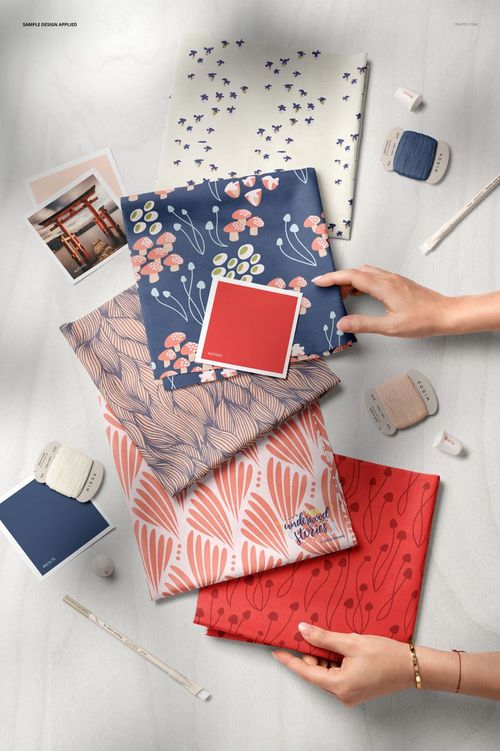 PSD mockup displaying five patterned fabric squares, color sample cards, thread spools, and hands arranging items on a pale background.