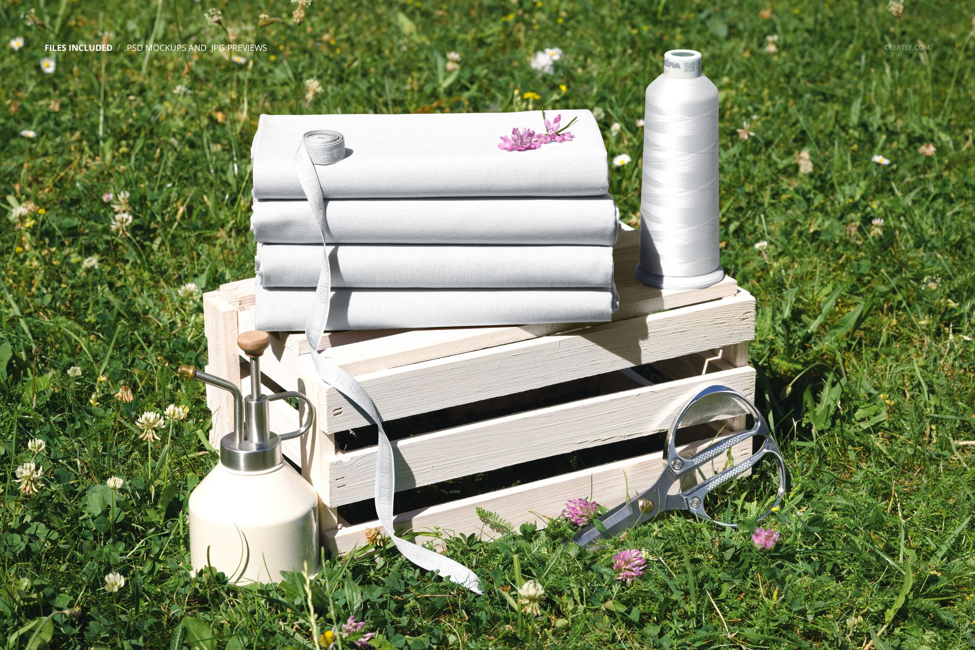 PSD mockup showing folded white jersey fabrics with sewing accessories on a crate, surrounded by green grass.