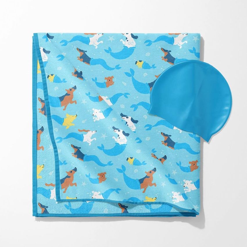 Beach towel mockup featuring a playful pattern of cartoon dogs as mermaids in blue tones, shown with a blue swim cap on a white background.