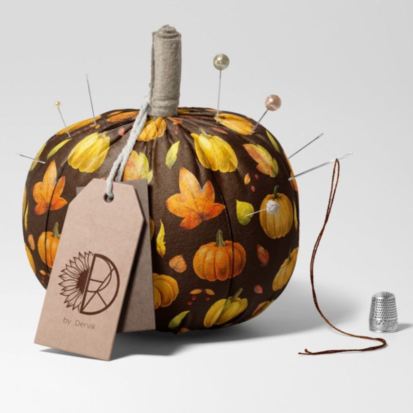 Fabric pumpkin mockup decorated with autumn leaves and pumpkin patterns, used as a pincushion with pins and a thimble nearby.