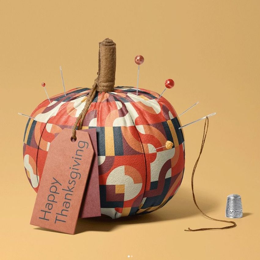 Fabric pumpkin mockup with a geometric orange and navy pattern, pins inserted, a brown tag reading 'Happy Thanksgiving', and a thimble on a beige background.