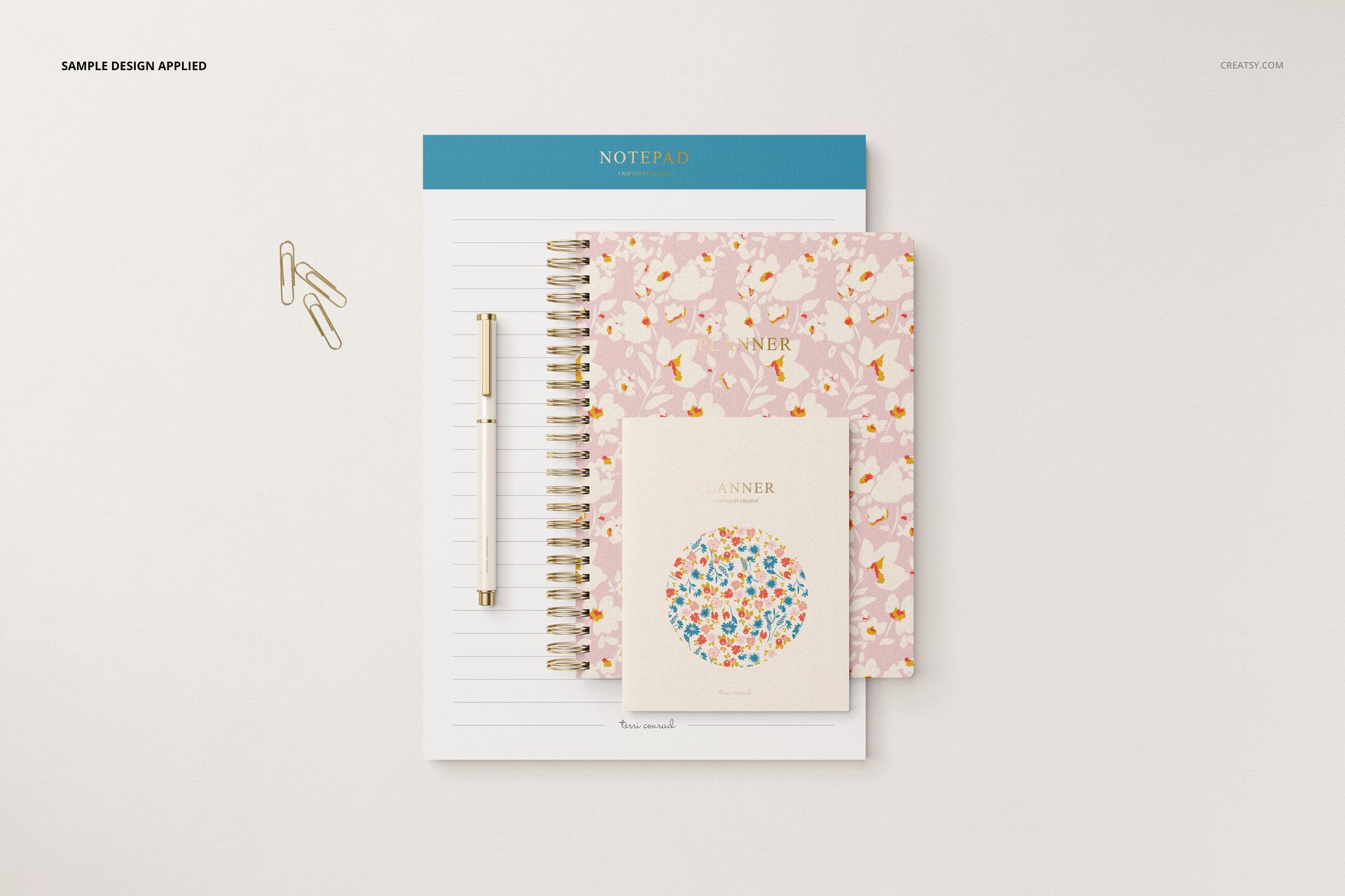 Stationery set mockup featuring a notepad, two floral-patterned planners, a pen, and paper clips on a light surface.