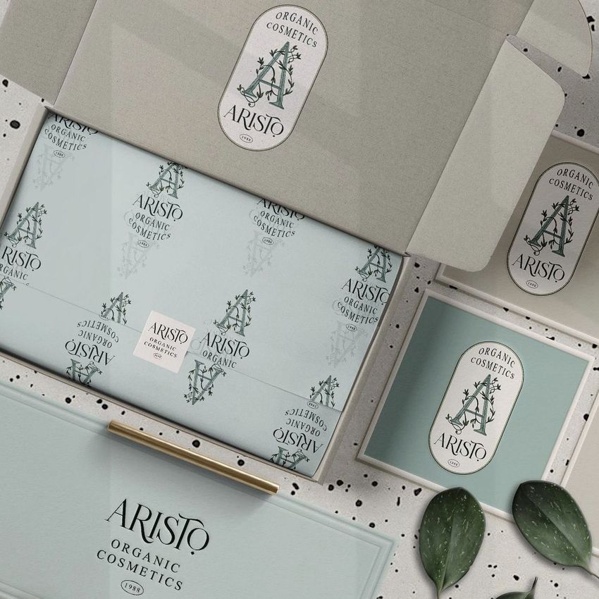 Mailing box mockup featuring an open cardboard box with custom printed tissue paper, branded cards, and green leaves on a speckled surface.