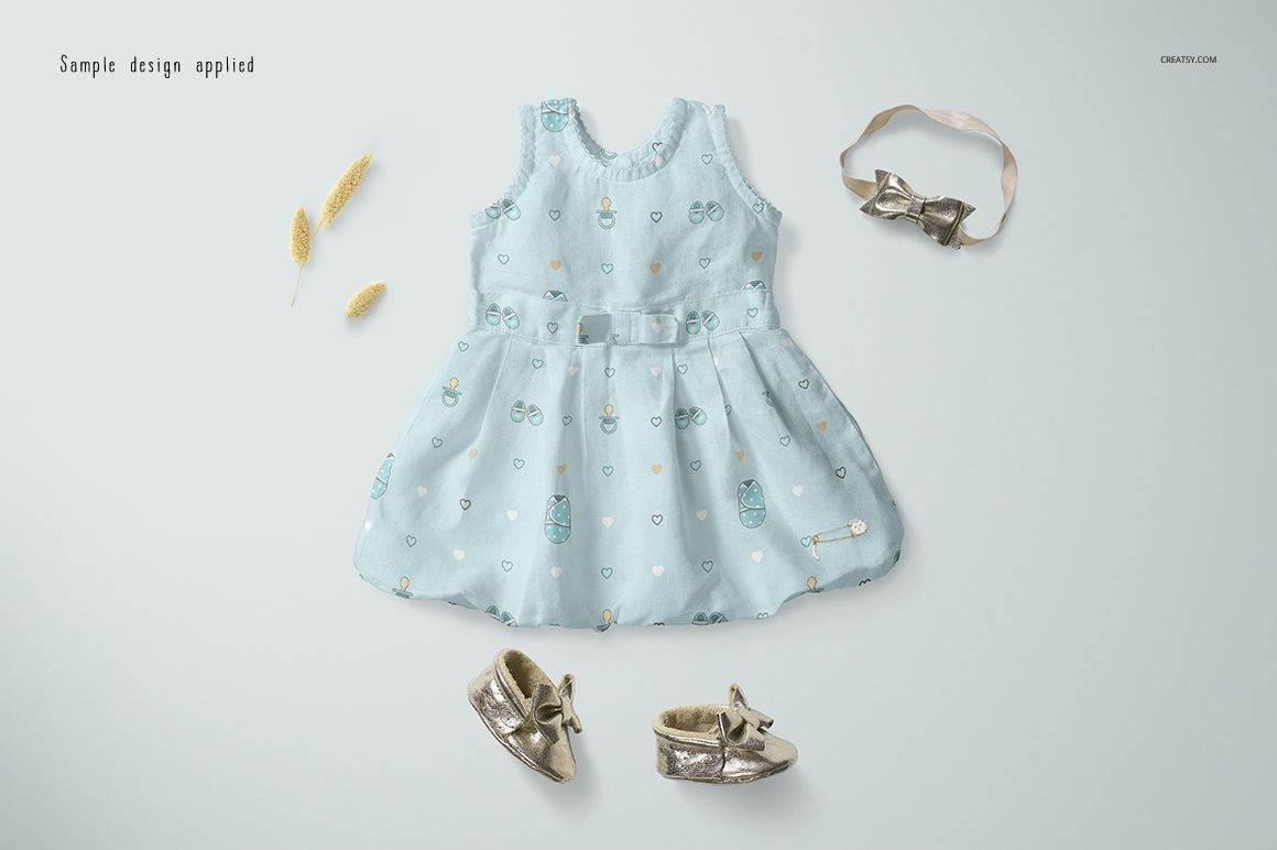 Baby dress mockup shown from top view, featuring a pale blue color and playful motifs, with gold shoes and headband nearby.