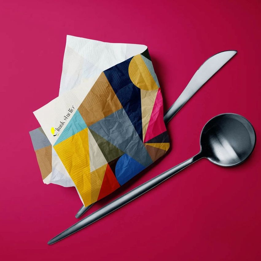 Paper napkins mockup featuring a crumpled napkin with a colorful geometric pattern, placed beside a spoon and knife on a magenta background.