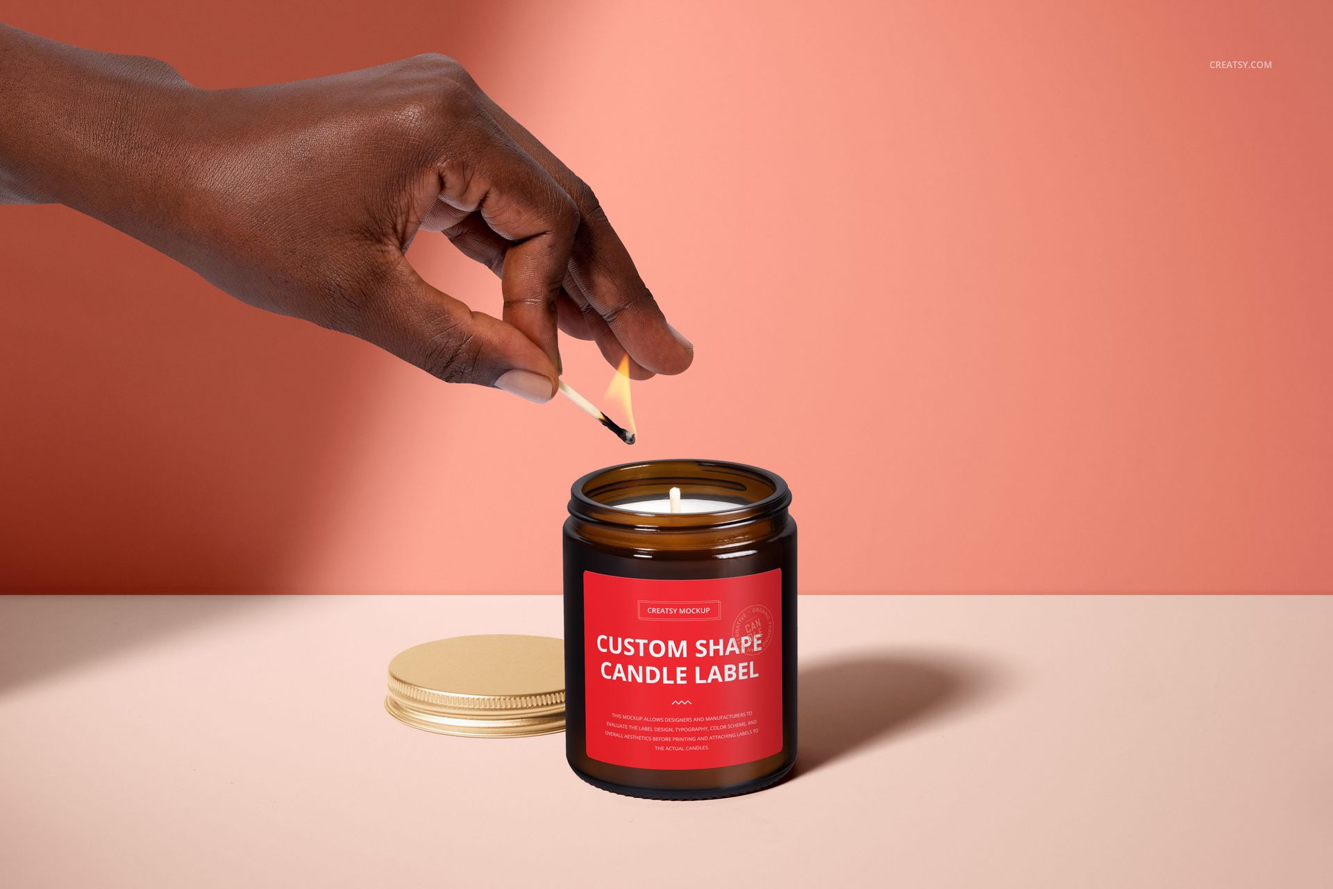 PSD mockup of amber glass candle jar with red label, cotton wick being lit, gold lid on table, minimal background.