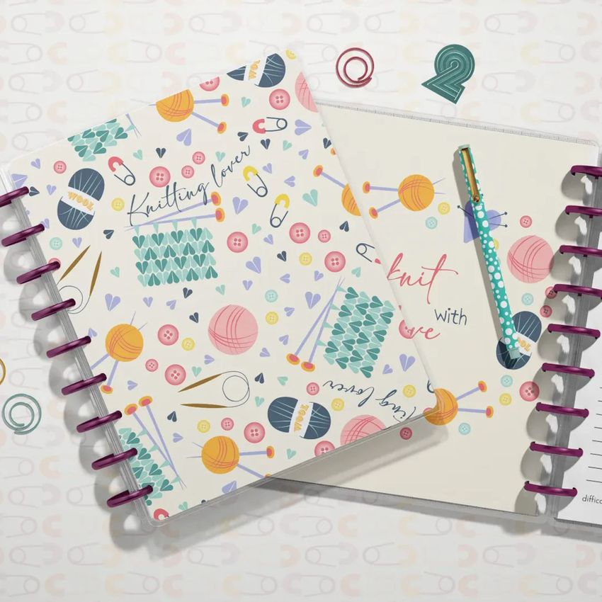 Top view of two spiral planners with knitting-themed covers, colorful yarn and needles illustrations, and a pen on white background.
