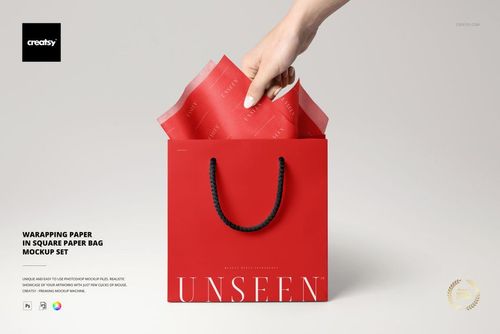 Hand holding red wrapping tissue paper above a square red gift bag with black handles on a light background.