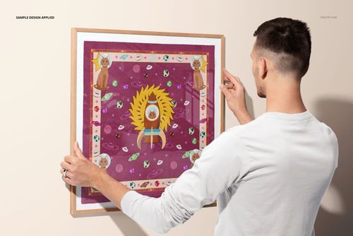 Person hanging a square picture frame with a silk scarf mockup featuring a colorful abstract pattern on a wall.