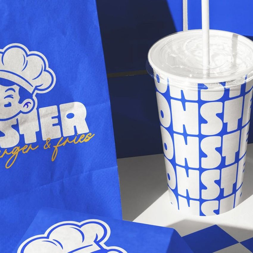 Burger branding mockup showing a blue paper bag, takeaway box, and a cup with bold white and blue patterns on a blue background.