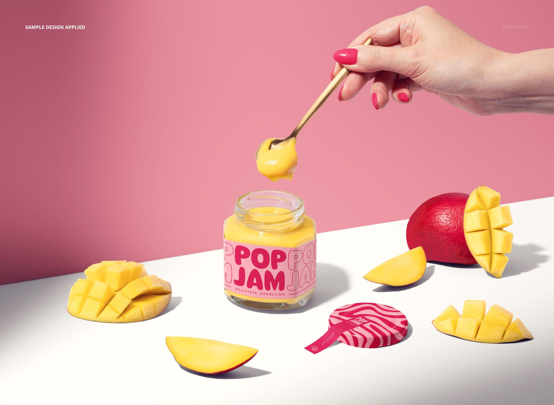 PSD mockup of a glass jam jar with yellow contents, mango pieces, and a hand scooping jam on a pink surface.
