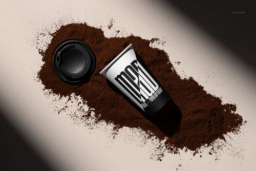 PSD mockup of a paper cup and lid resting on scattered ground coffee, showing a modern black and white design.