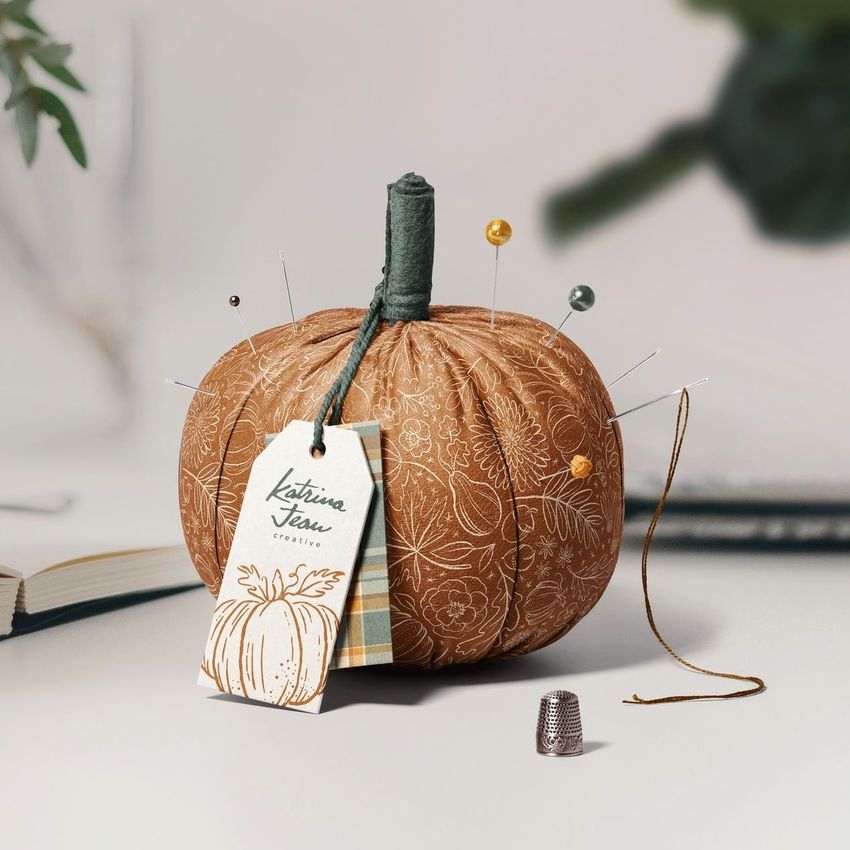 Fabric pumpkin mockup displayed on a light surface, decorated with sewing pins, a thread, and a paper tag attached.