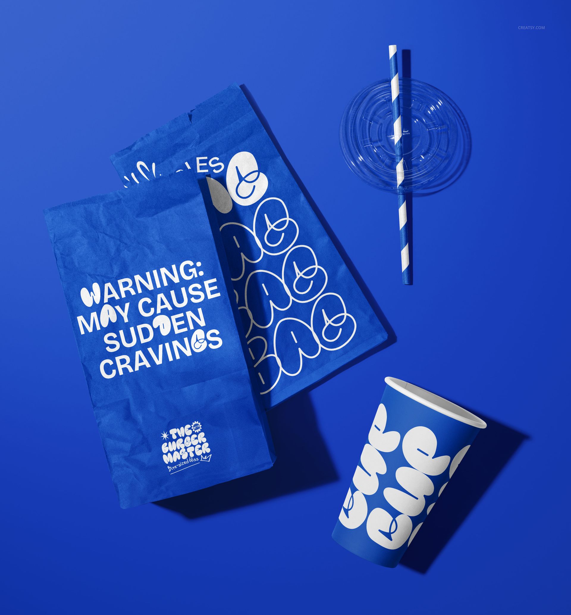 PSD mockup showing two blue paper bags and a takeaway cup with white graphic designs, arranged on a blue background.
