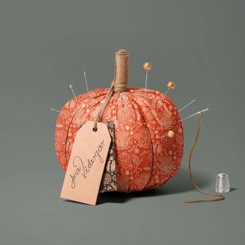 Fabric pumpkin mockup with orange patterned fabric, sewing pins, a brown string, and a thimble on a plain gray background.