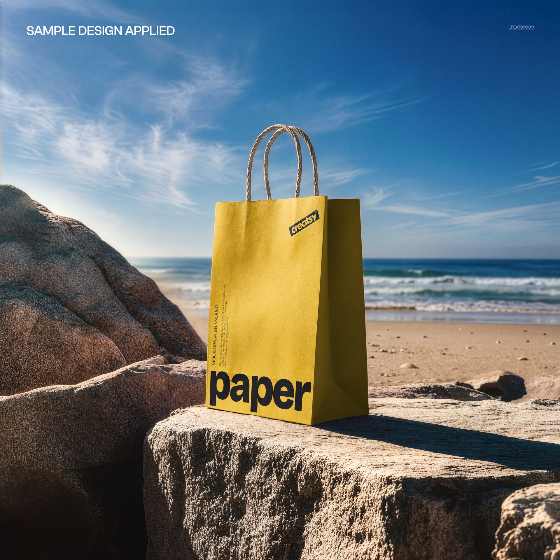 Front view of a yellow paper shopping bag mockup on a beach, with sunlight, rocks, and waves in the background.