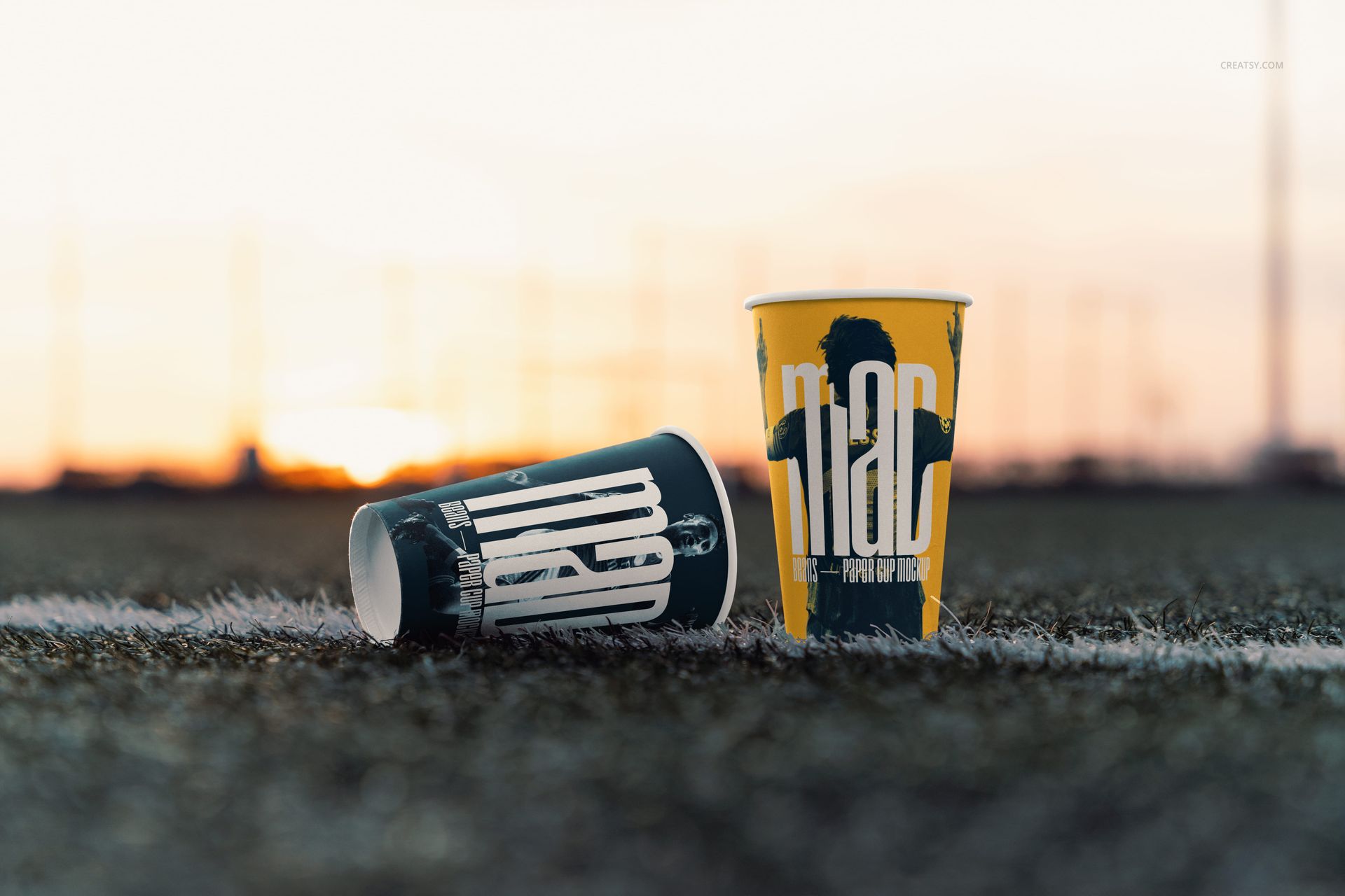 PSD mockup featuring paper cups with artistic prints, positioned on grass with blurred stadium lights in the background.
