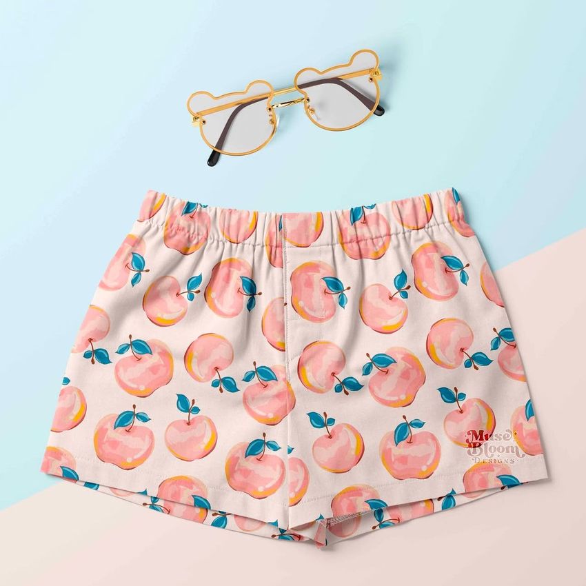 Kids shorts mockup featuring a playful apple pattern in pink and blue, displayed flat on a pastel background with bear-shaped glasses.