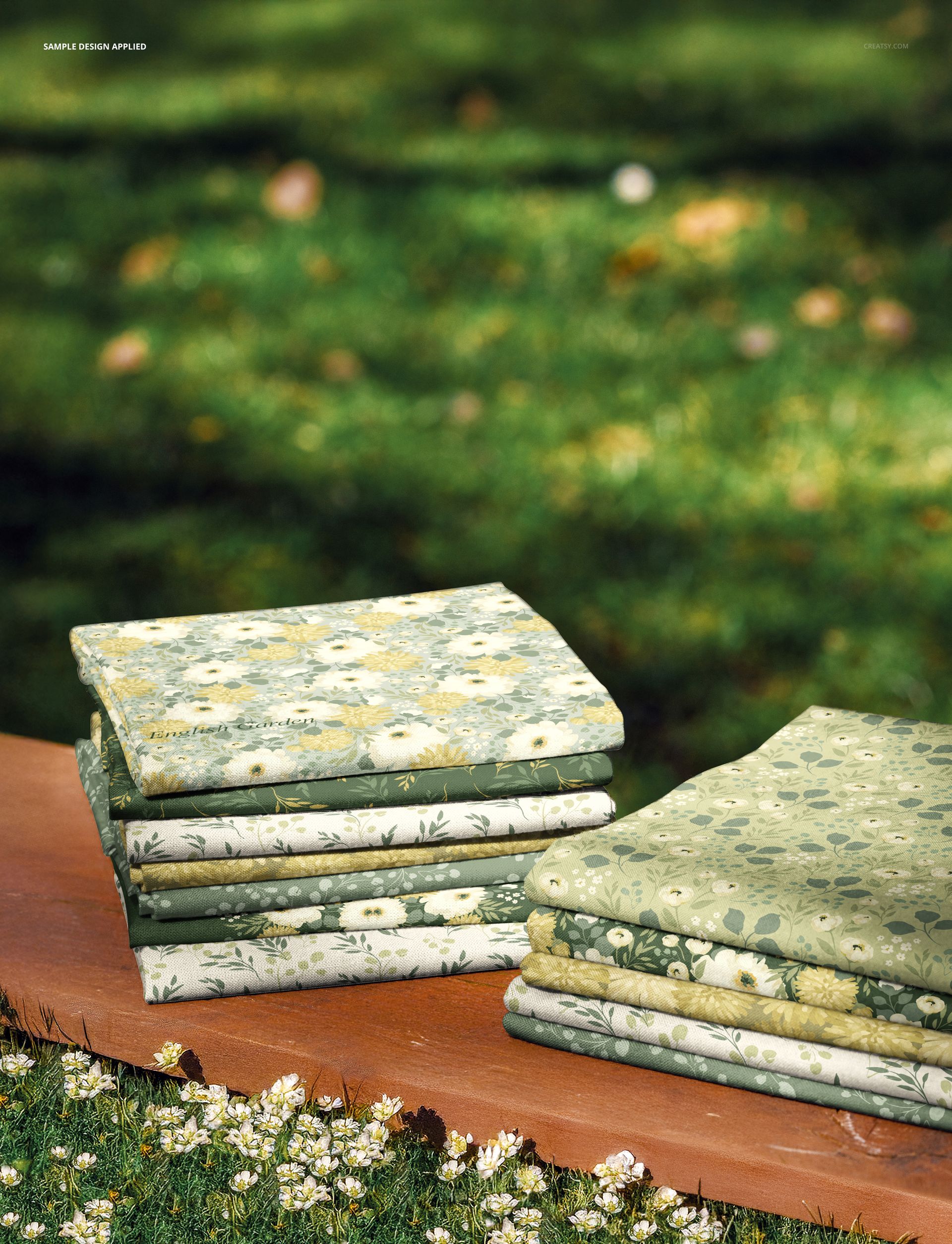 PSD mockup showing multiple fabric pieces with botanical prints stacked on a wooden plank in a meadow scene.