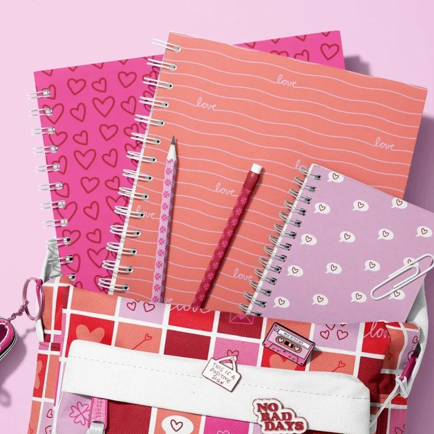 School backpack mockup with spiral notebooks in pink and red tones, decorated with hearts and text, on a pastel background.