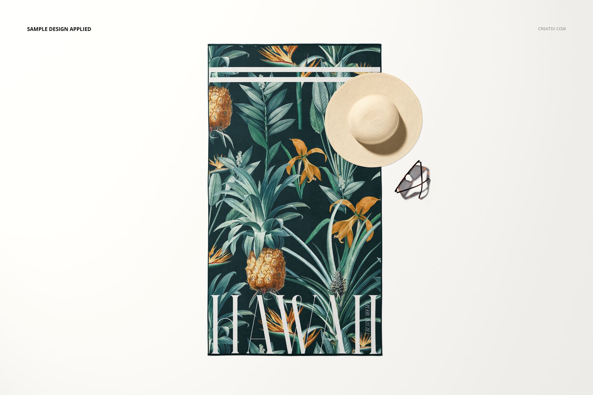 PSD mockup showing a beach towel with vibrant tropical print, laid flat with a beige sun hat and sunglasses on a plain backdrop.