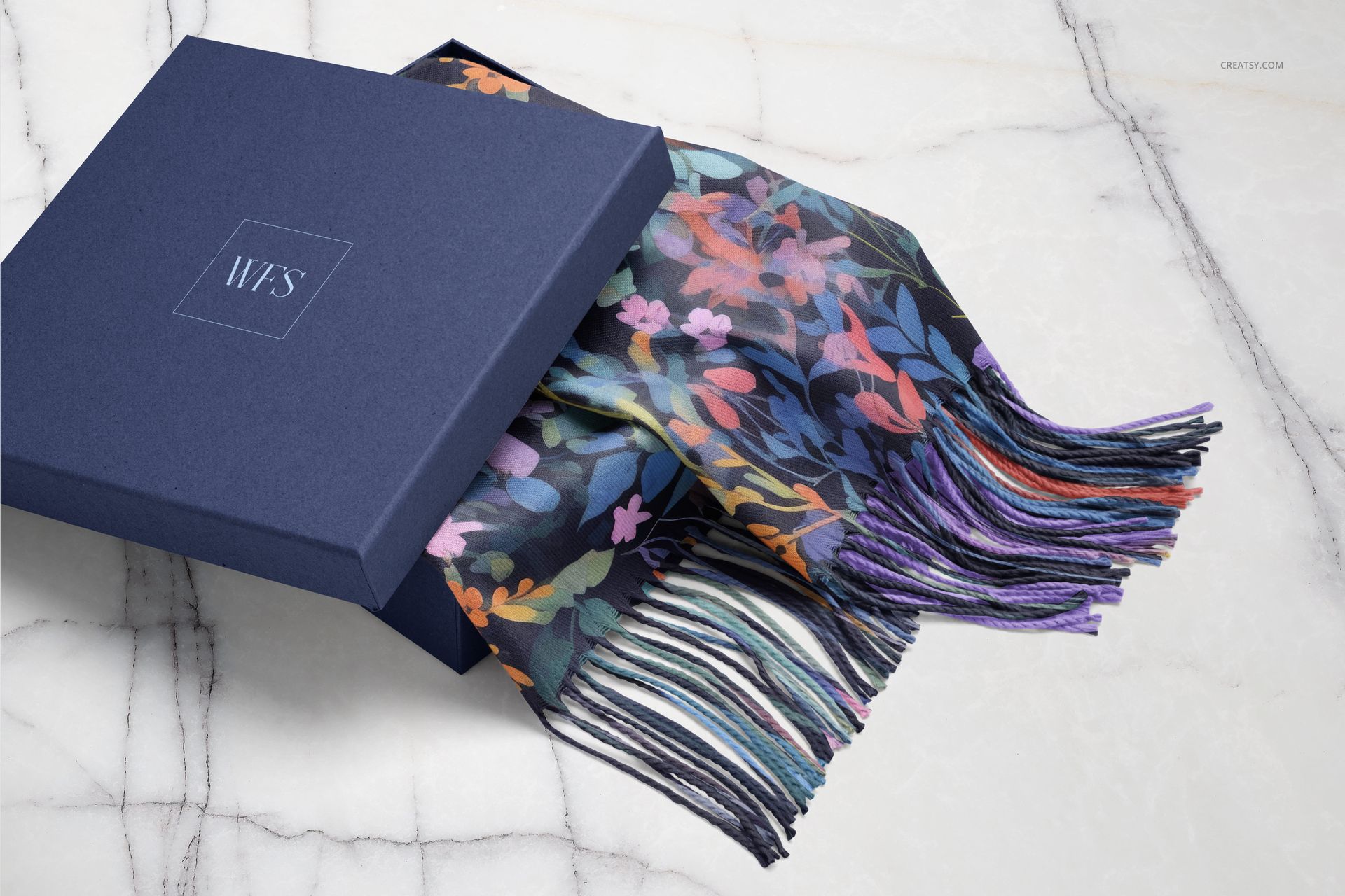 Woven fringed scarf with colorful floral pattern partially placed inside a dark blue box on a marble surface.