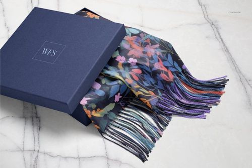 Woven fringed scarf with colorful floral pattern partially placed inside a dark blue box on a marble surface.
