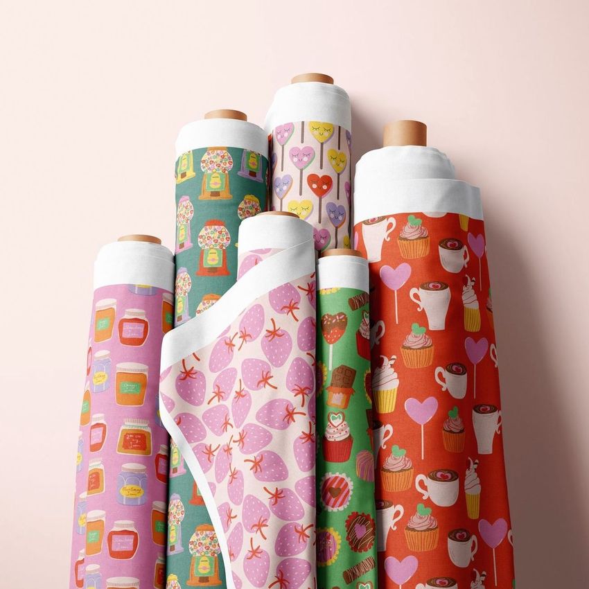 Fabric rolls mockup featuring seven upright rolls with colorful patterns, including sweets, jars, and hearts, on a light pink background.