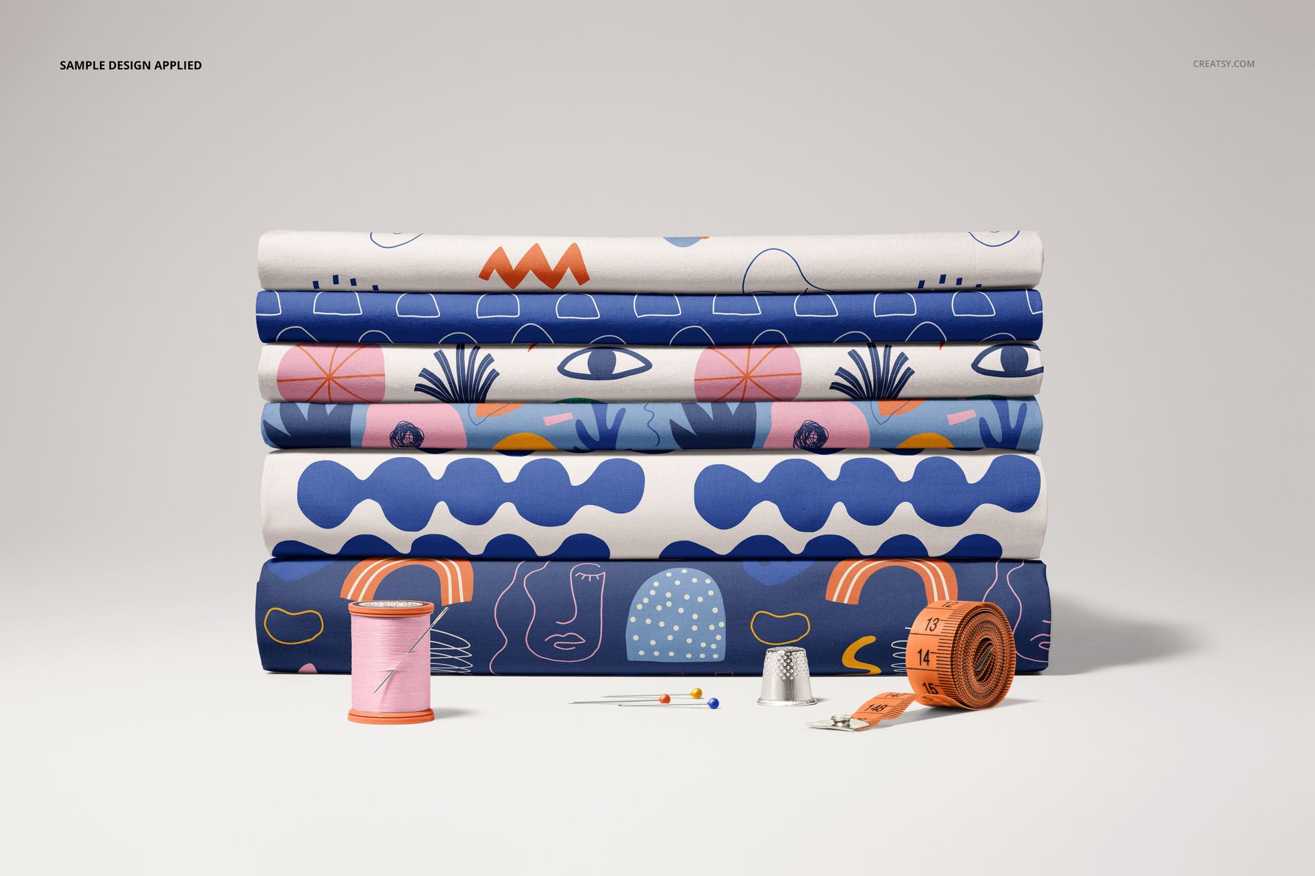 Folded fabric mockup featuring six stacked textiles with blue, pink, and orange designs, sewing tools in foreground.