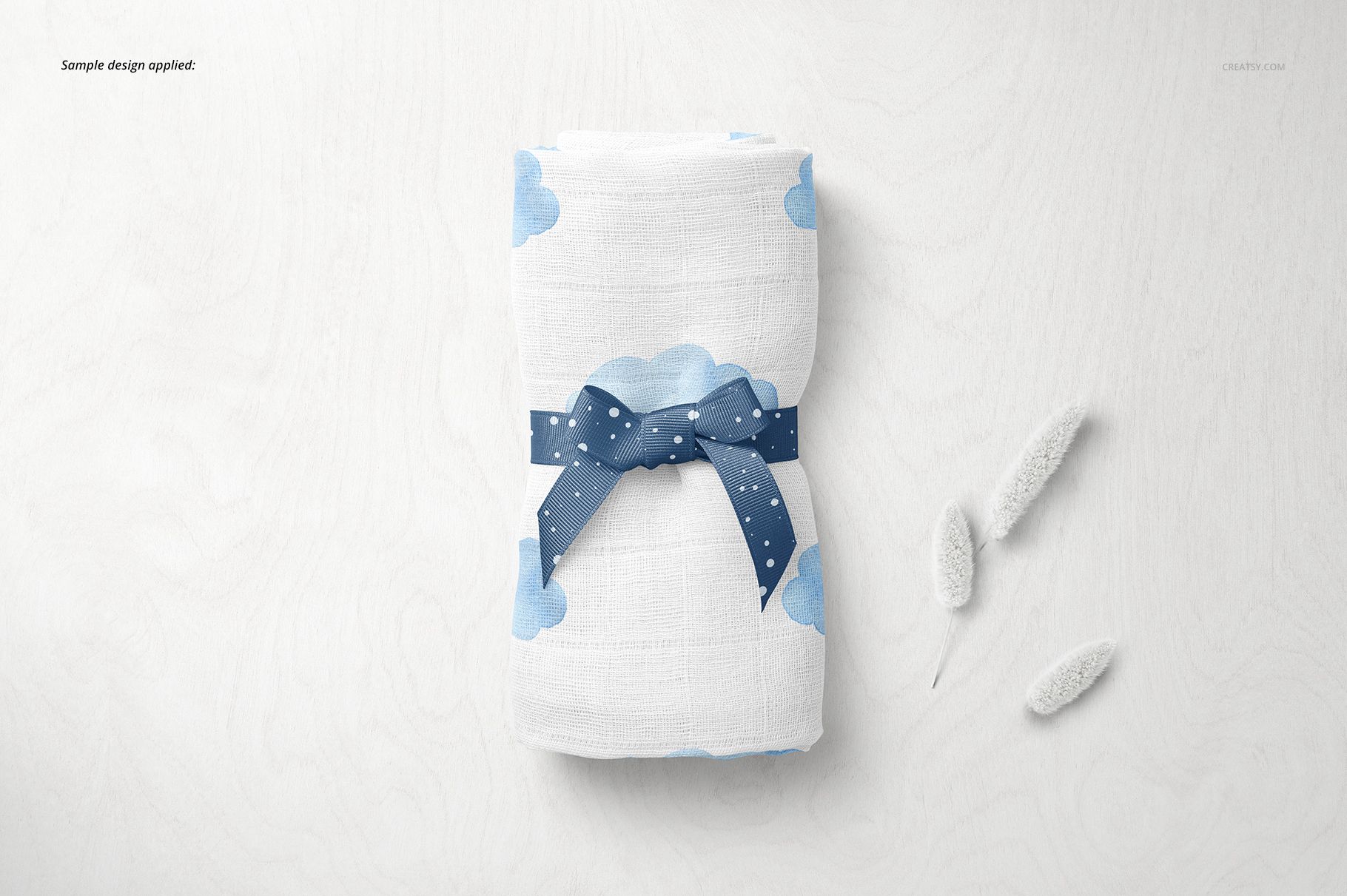 PSD mockup showing a folded baby muslin nappy with cloud design and ribbon, styled on a light tabletop with props.