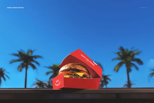 Red burger packaging mockup featuring an open box with a burger, placed outdoors with palm trees and clear sky.