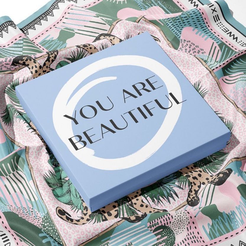 Silk scarf box mockup featuring a square blue box with bold text on the lid, placed on a patterned silk scarf with animal motifs.