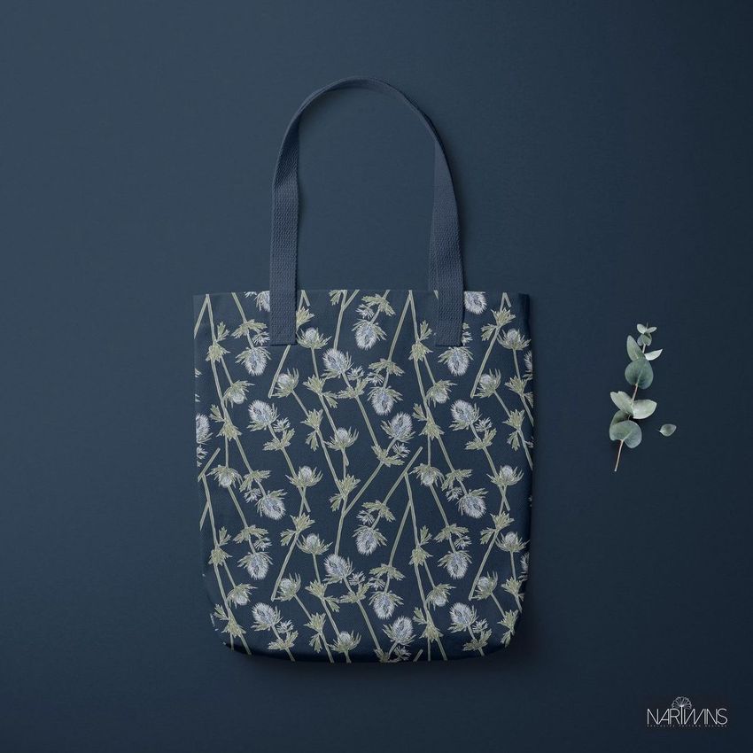 Polyester tote bag mockup featuring a floral pattern in muted tones, displayed flat on a dark blue background with a sprig of greenery.
