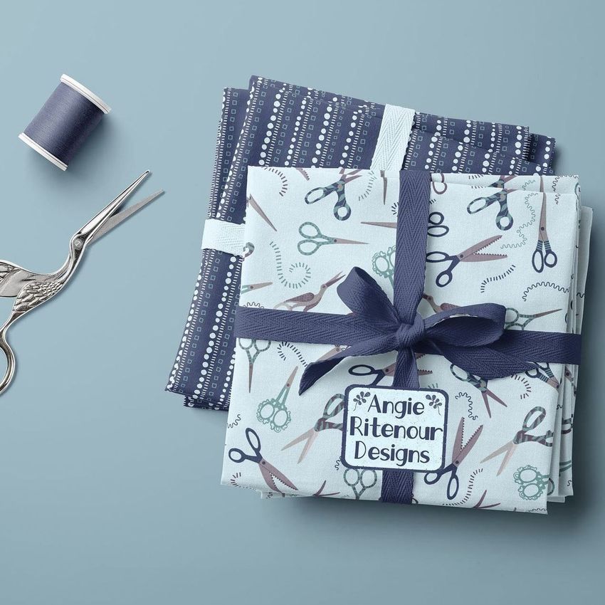 Fabric stack mockup showing two folded fabric pieces with sewing-themed patterns, ribbon, scissors, and thread on a blue background.
