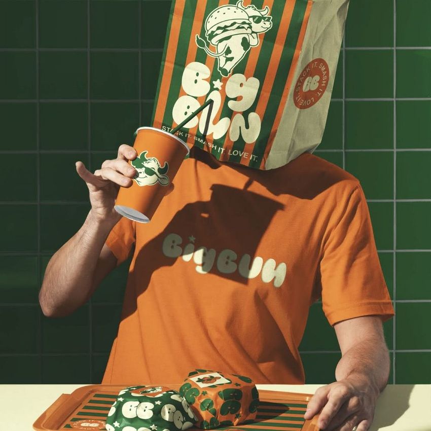 Fast food mockup scene featuring a man with a branded paper bag on his head, holding a cup and surrounded by wrapped food items.