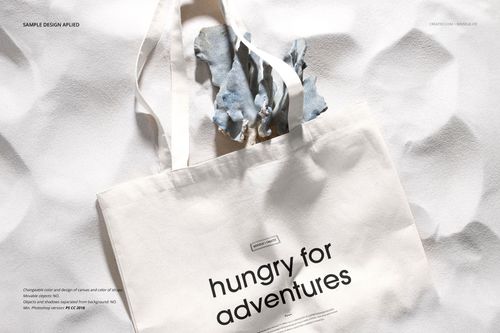 Front view of a white tote bag mockup with black text, placed on a textured white surface with fabric inside.