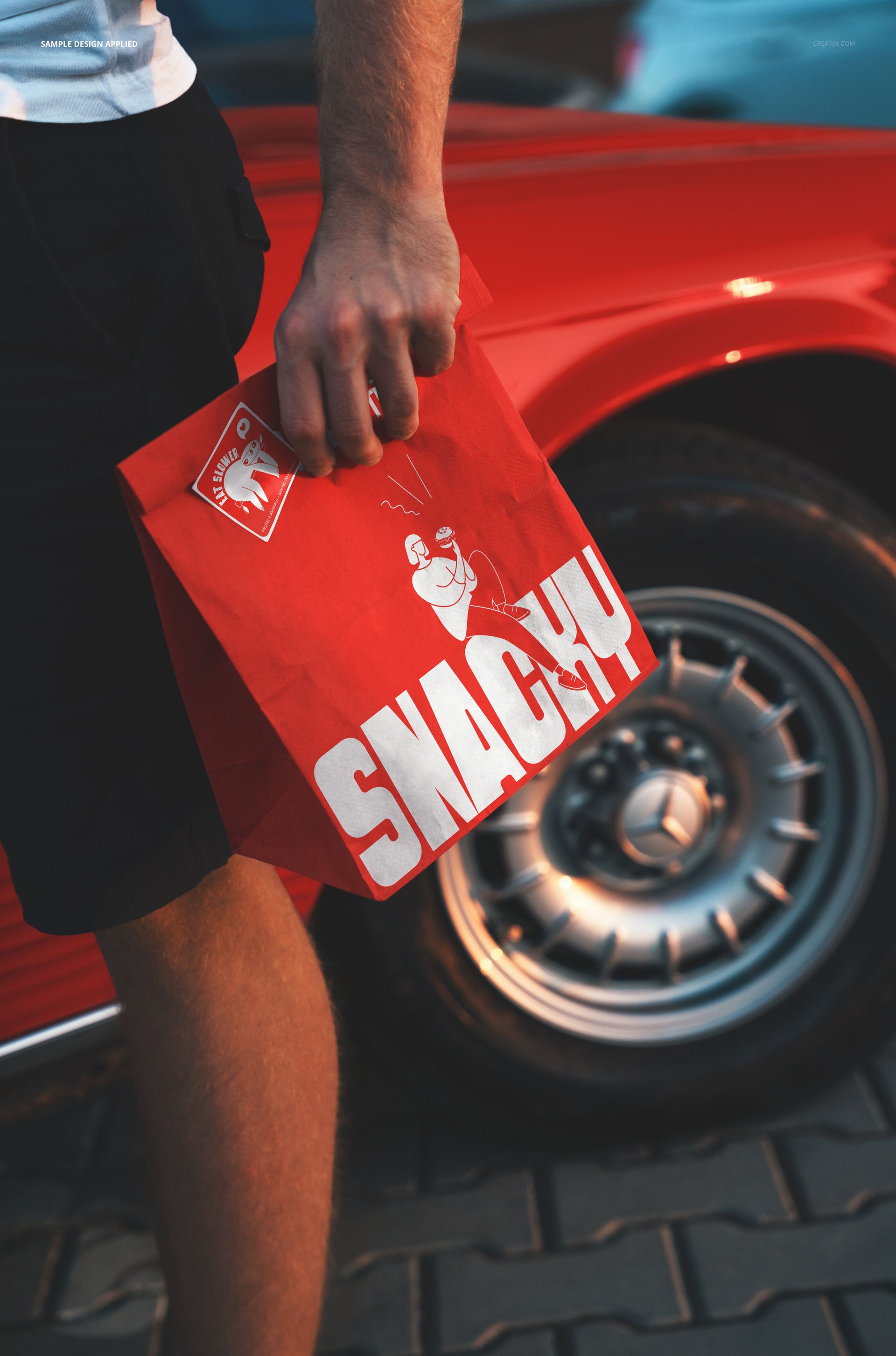 Close-up of a red paper bag mockup featuring white artwork, held beside a retro car parked on pavement.