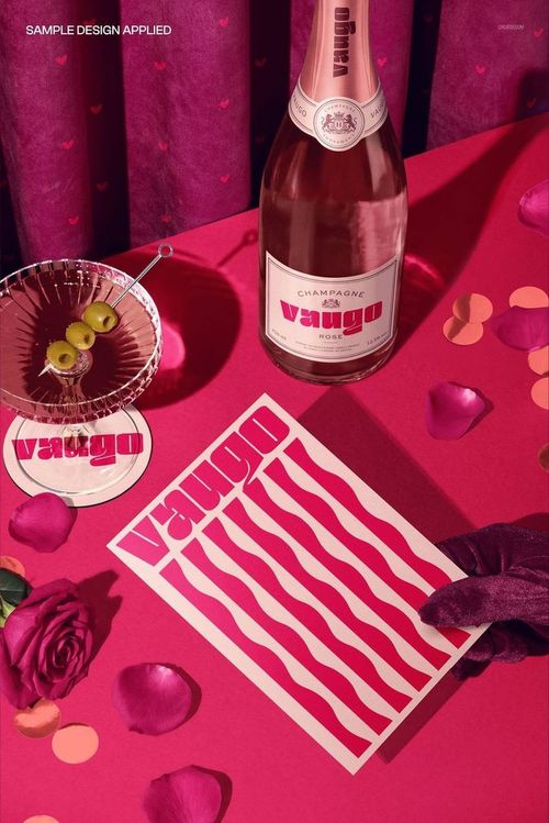 Valentine’s Day greeting card mockup on a pink table with a bottle of champagne, velvet curtain, and rose petals.