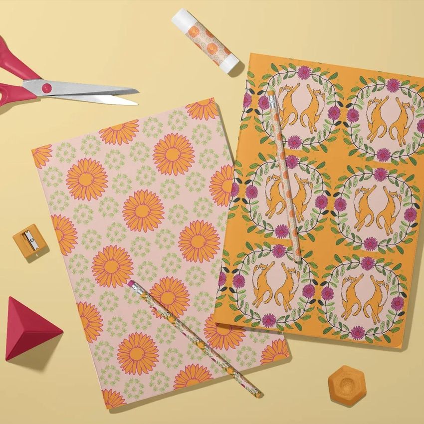 Stationery mockup featuring two classic A4 notebooks with colorful floral and cat patterns, arranged on a yellow surface with scissors, a pencil, a sharpener, and geometric props.