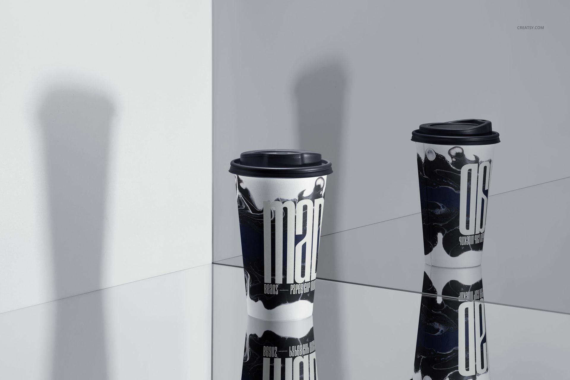 Two paper drinkware cups with black lids and artistic black-and-white designs, shown on a reflective tabletop in a mockup scene.