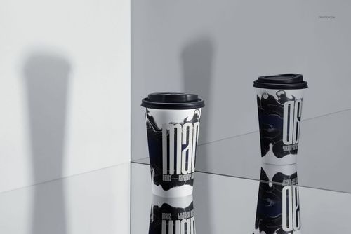 Two paper drinkware cups with black lids and artistic black-and-white designs, shown on a reflective tabletop in a mockup scene.