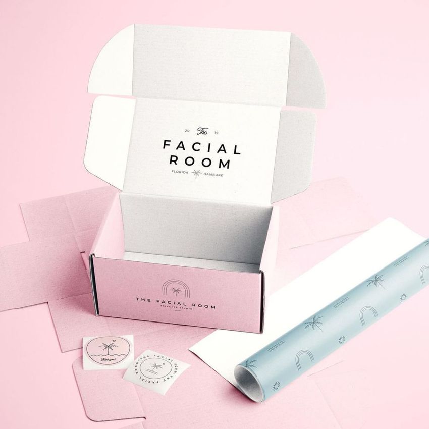 Mailing box mockup with pastel pink exterior and white interior, featuring printed text and logo, shown with branded tissue paper and stickers on a light pink background.