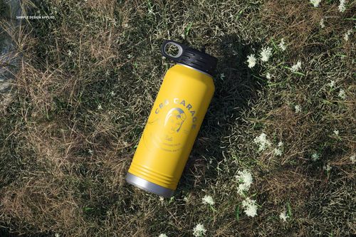 Top view of a yellow water bottle mockup with black cap and printed design, placed on grassy ground outdoors.