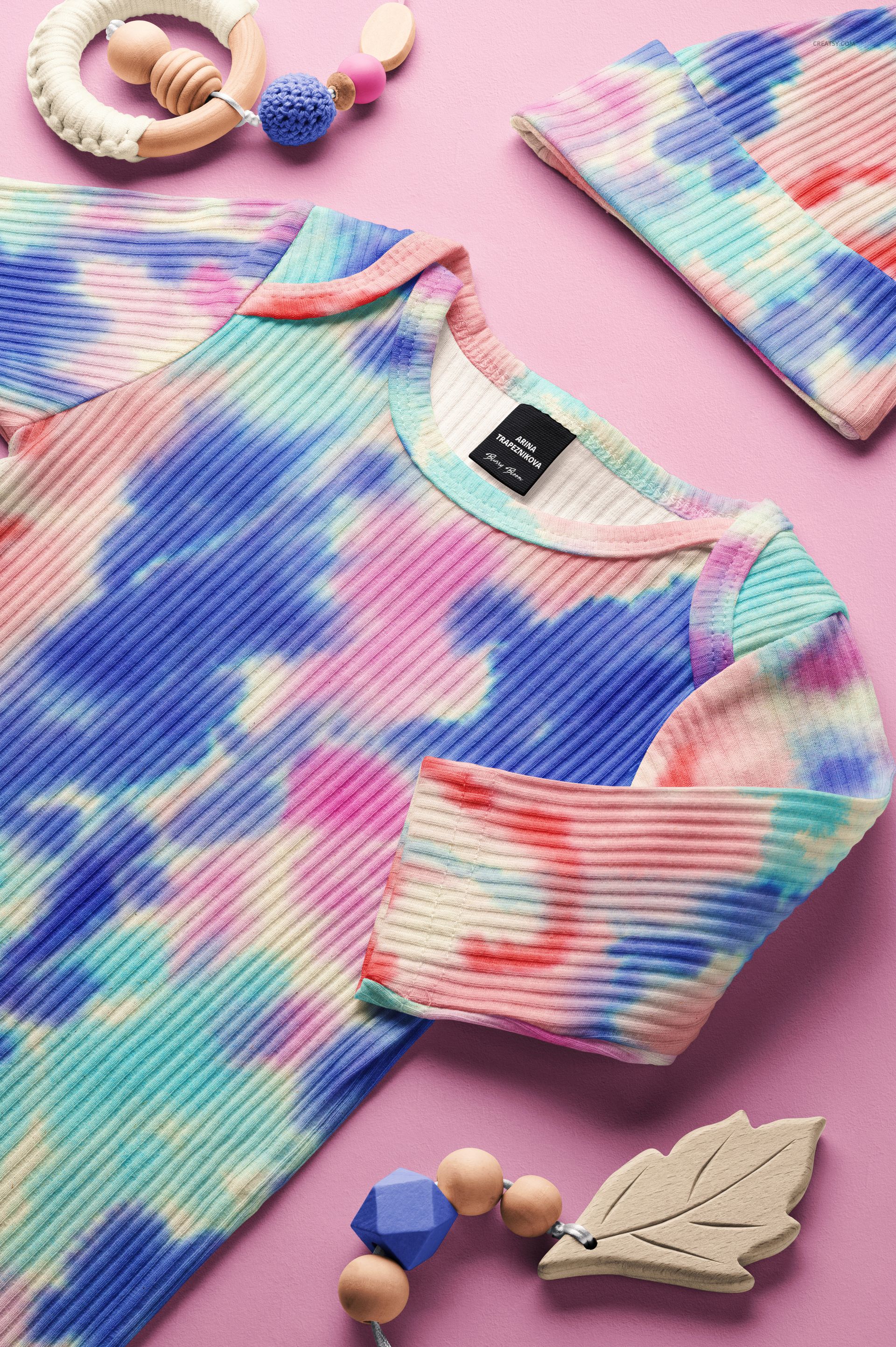 Baby ribbed gown and hat mockup with blue, pink, and purple tie-dye pattern, arranged on a pink surface.