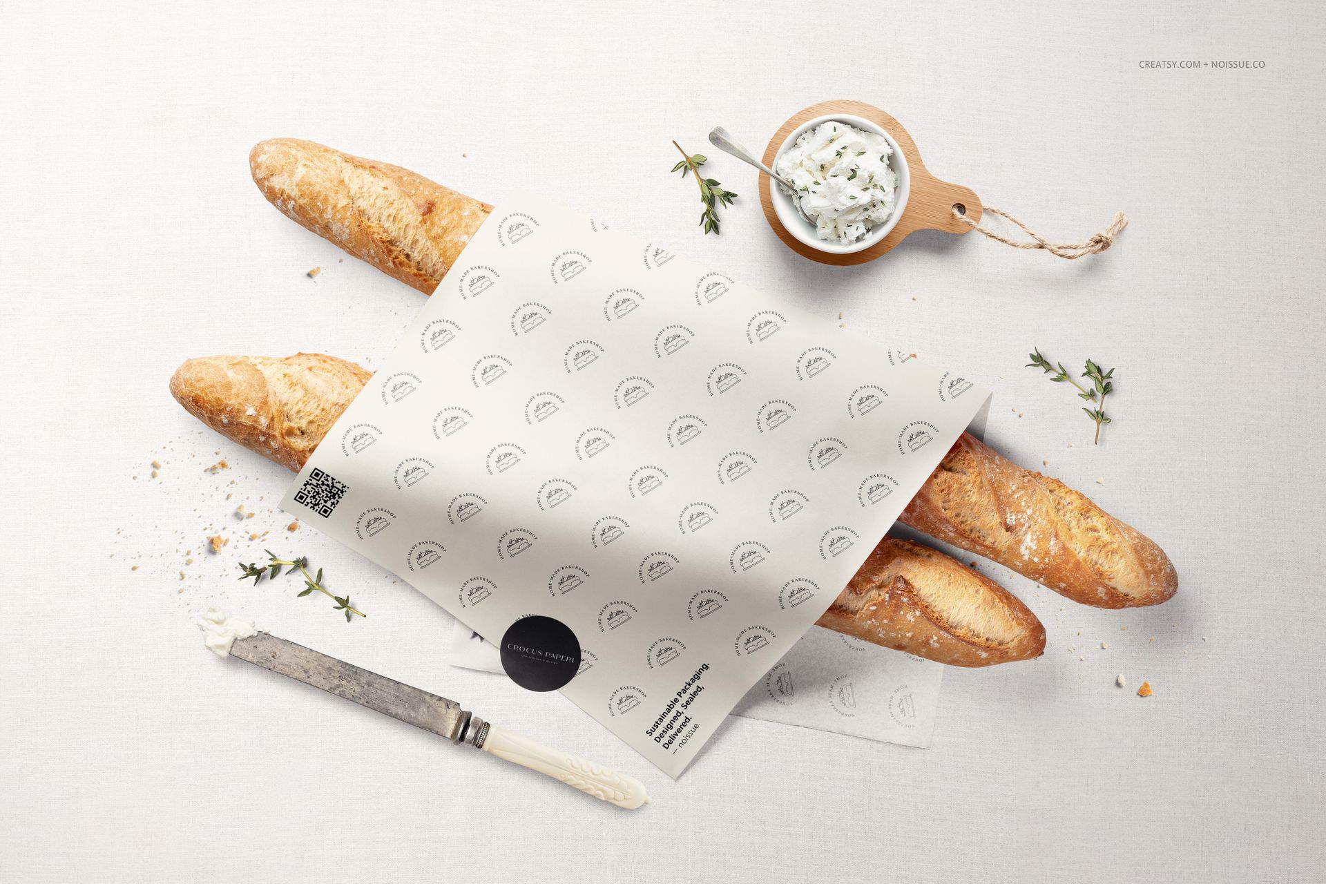 Top-down view of baguettes covered by printed food safe paper mockup, with a knife and dip bowl on a white cloth.