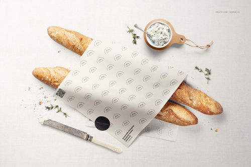 Top-down view of baguettes covered by printed food safe paper mockup, with a knife and dip bowl on a white cloth.