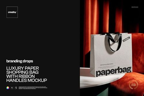 Front view of a paper shopping bag mockup featuring black ribbon handles, positioned beside a white round table and red drapes.