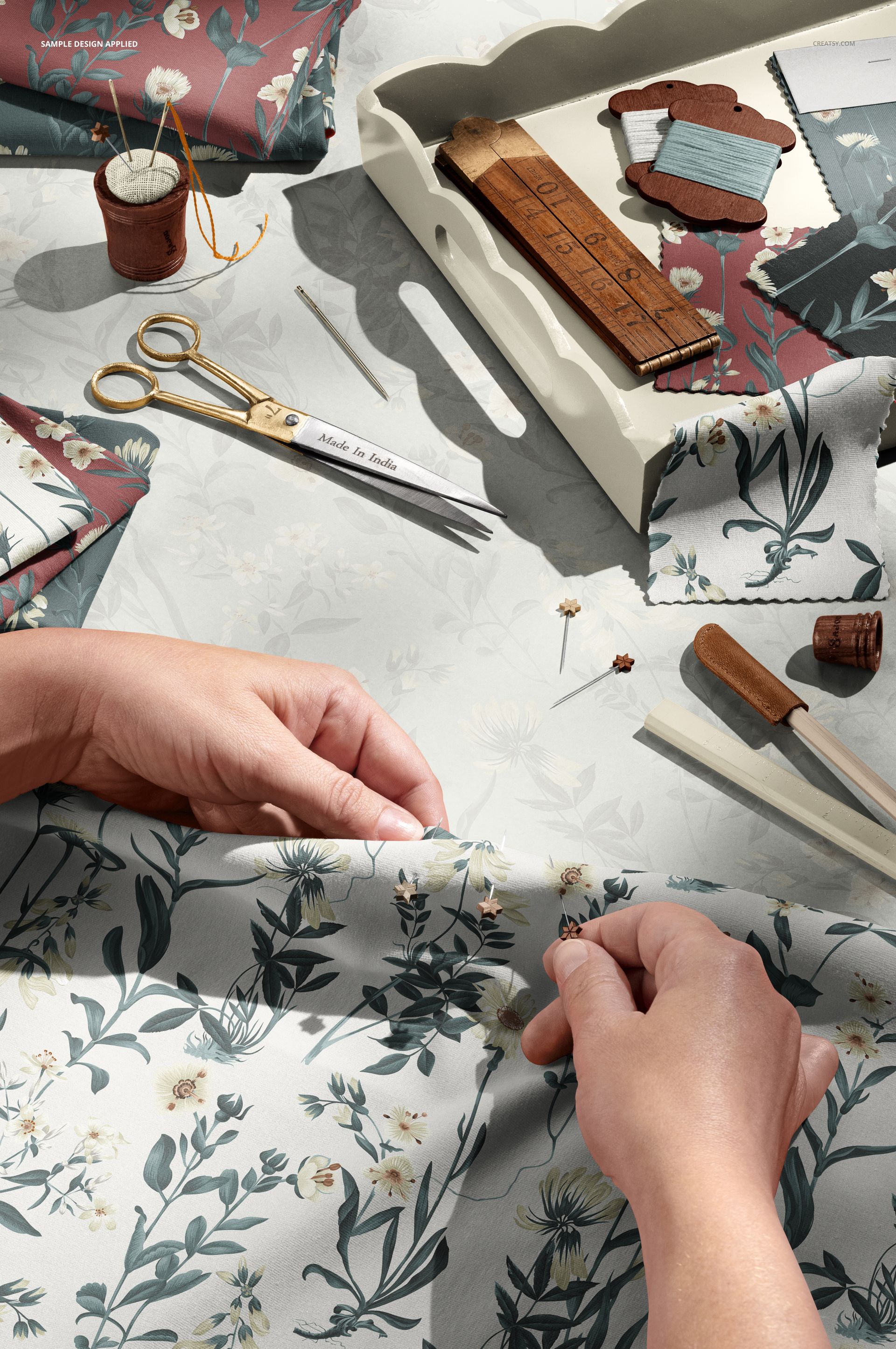 Close-up view of tailor hands working with flower-patterned jersey fabric, surrounded by sewing accessories and mockup props.
