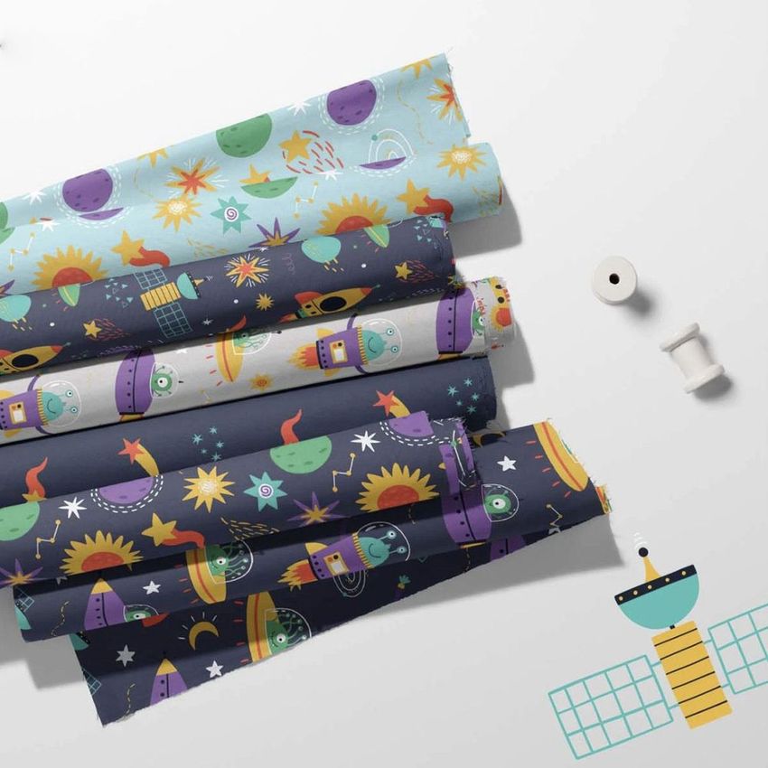 Fabric rolls mockup featuring colorful space-themed patterns with planets, rockets, and stars on a light surface with sewing props.