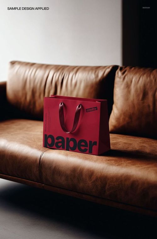 PSD mockup of a red paper shopping bag with black lettering, displayed on a tan leather sofa in soft lighting.